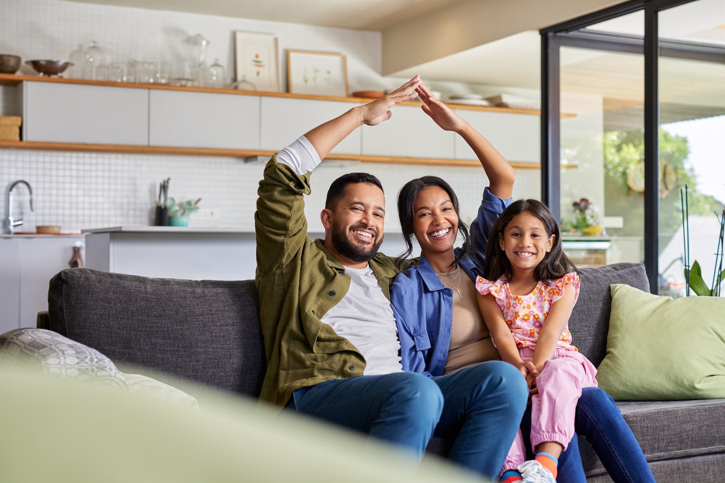 Happy man, woman, and child sitting together on couch making an outline of a house with their arms
